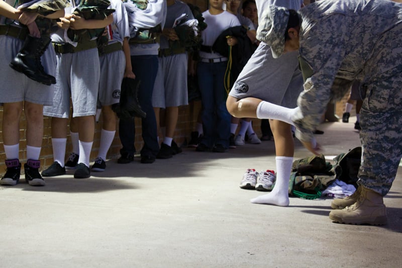 Female cadets line up for intake. A female drill instructor looks for contraband by inspecting pockets, socks and shoes, and performs a final pat-down before allowing cadets in the building.