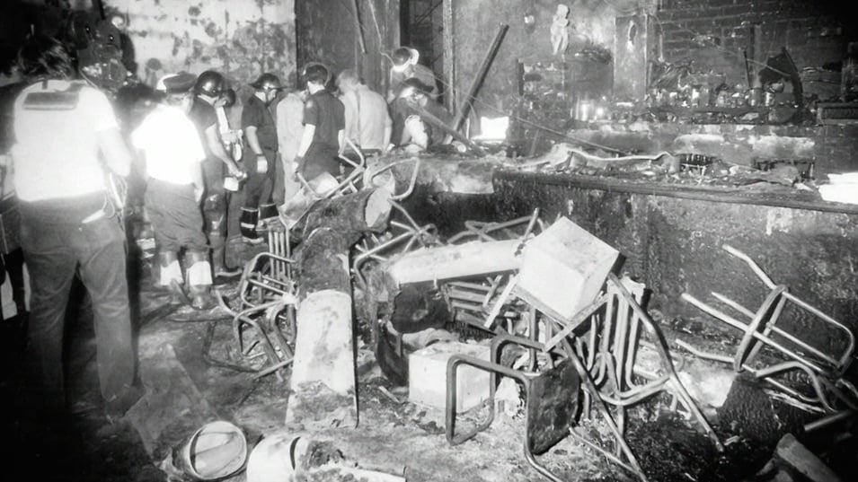 A black-and-white image of the aftermath of the UpStairs Lounge fire, with firefighters and upturned barstools in a charred room.