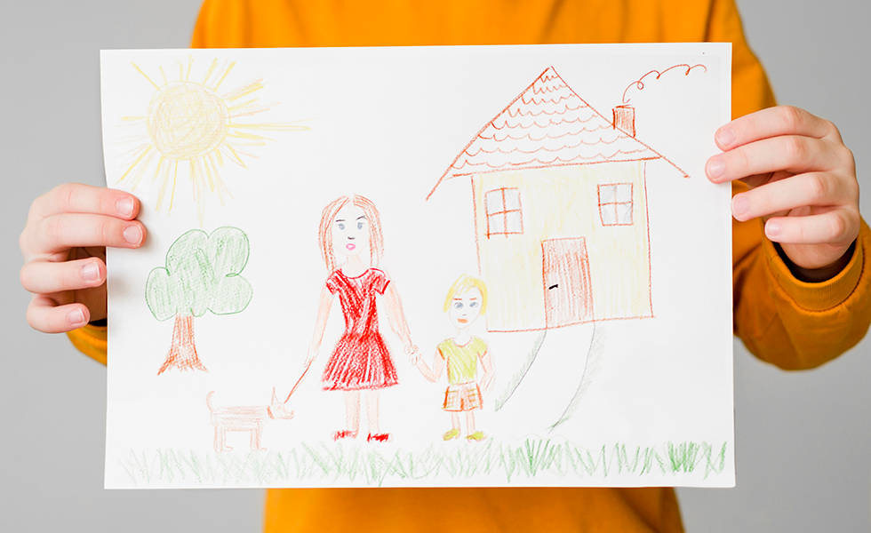 A person holds up a drawing of a mother and child in front of a house.