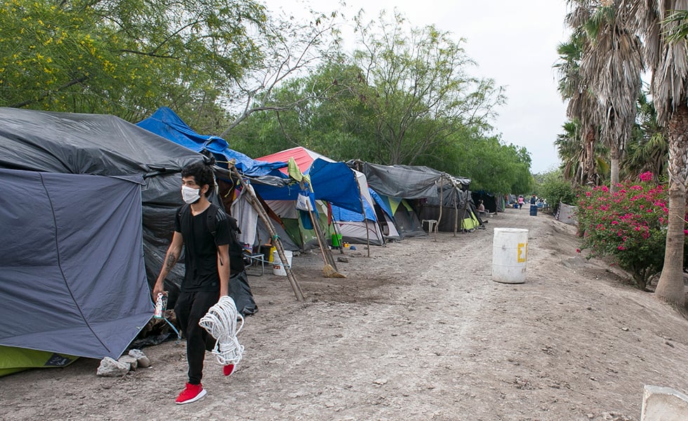 En esta foto del martes 14 de abril de 2020, un joven con una mascarilla camina por el campamento de migrantes durante la pandemia de coronavirus en las afueras de El Puente Nuevo en Matamoros, México. (Denise Cathey / The Brownsville Herald vía AP)