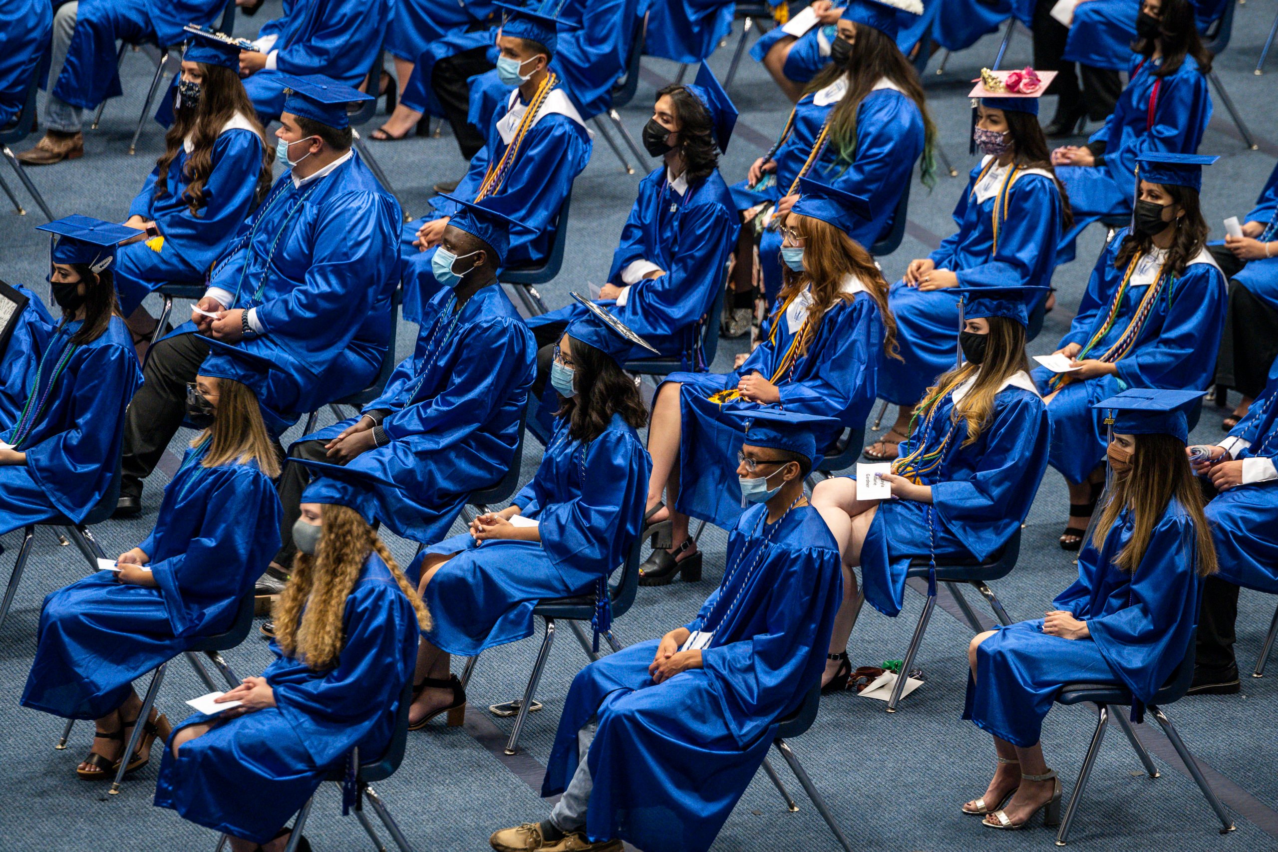 Students in a seeming ocean of blue gowns and caps listen, seated in rows of plastic chairs at a graduation ceremony.