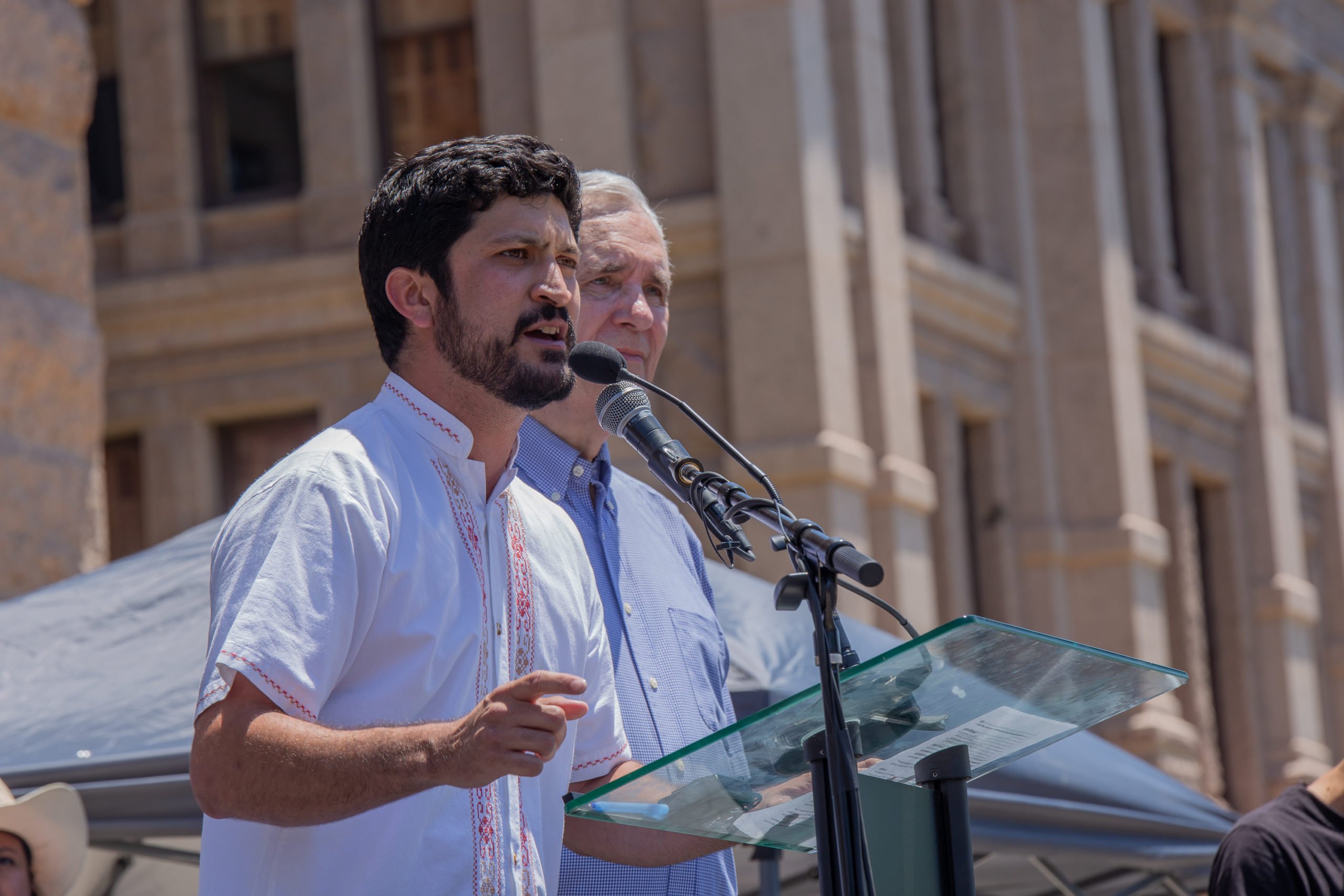 Greg Casar, wearing a white button-down shirt, speaks at a podium while standing next to Lloyd Doggett (who is wearing a blue button-down shirt).