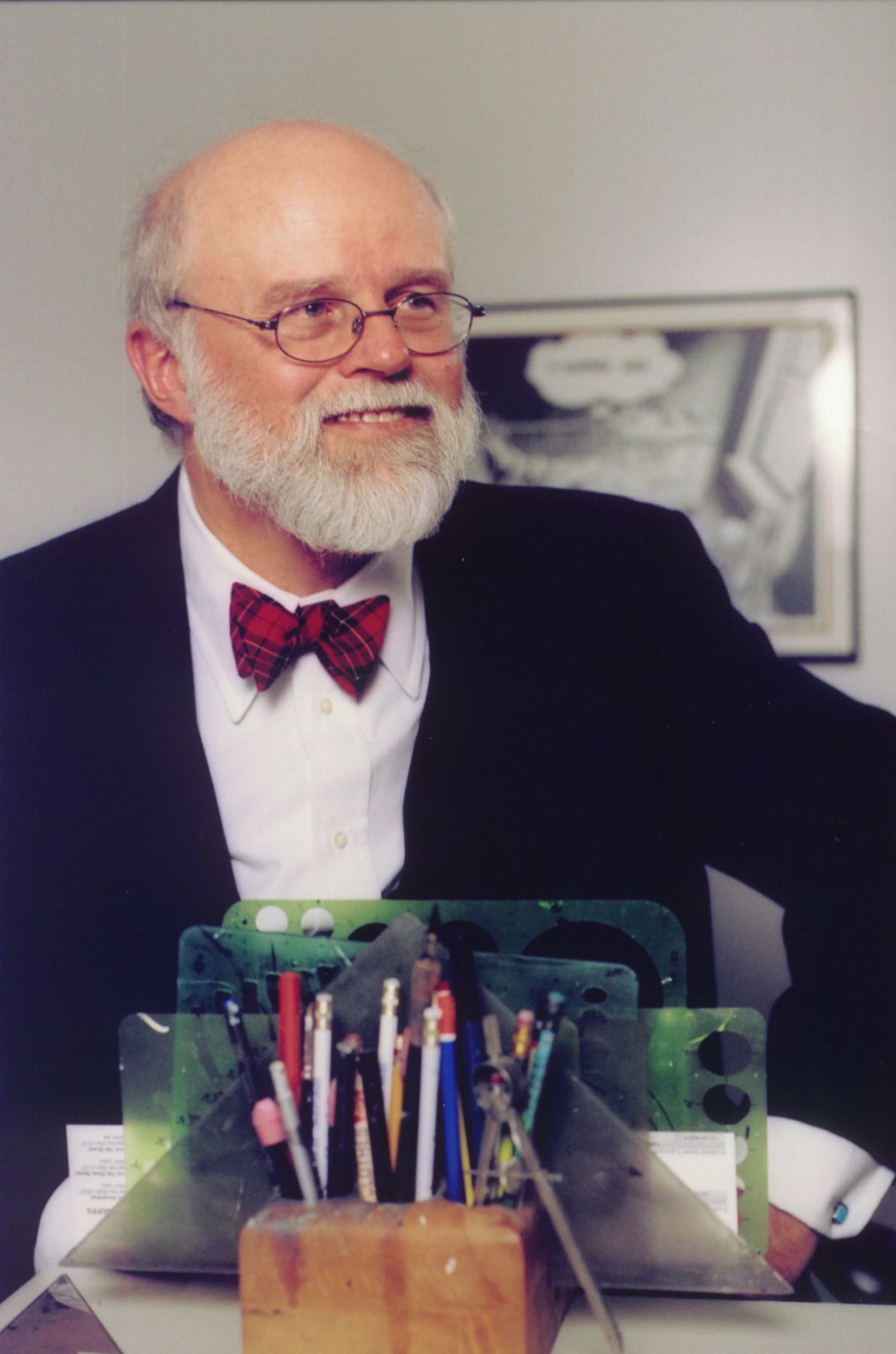 Ben Sargent, a bald, older white man with a bushy white beard is dressed in a suit and red bowtie, with his artistic tools, including pens, in front of him.