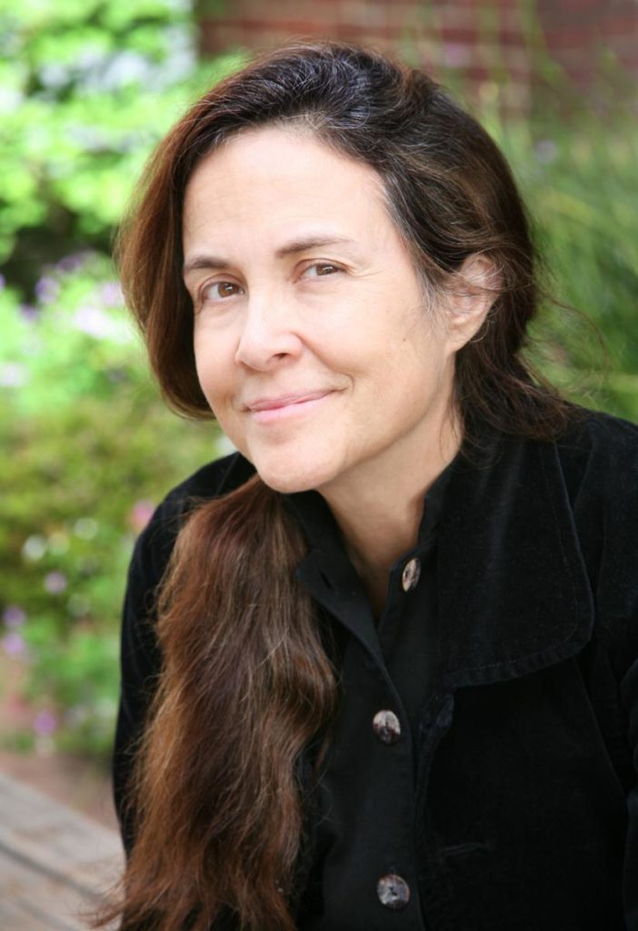 Naomi Shihab Nye has her brown hair in a pony tail hanging over her right shoulder. She's wearing a black collared button-down shirt and standing outdoors in a garden environment.