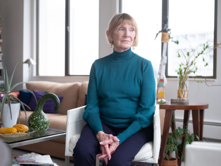 Wanda sits with her hands folded in her lap, looking to the right with a serious expression. She is a white woman with chin-length red-blonde hair. She's surrounded in her living room by plants and decorative gourds.