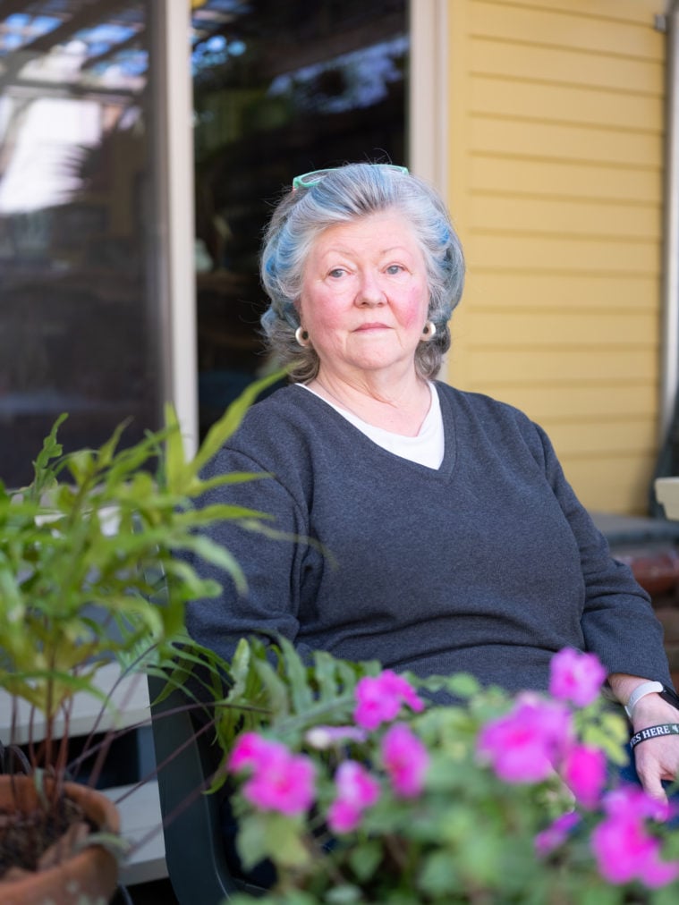 A rosy-cheeked white woman with small hoop earrings and permed gray hair sits on her porch, behind a pair of potted plants. One is blooming pink.