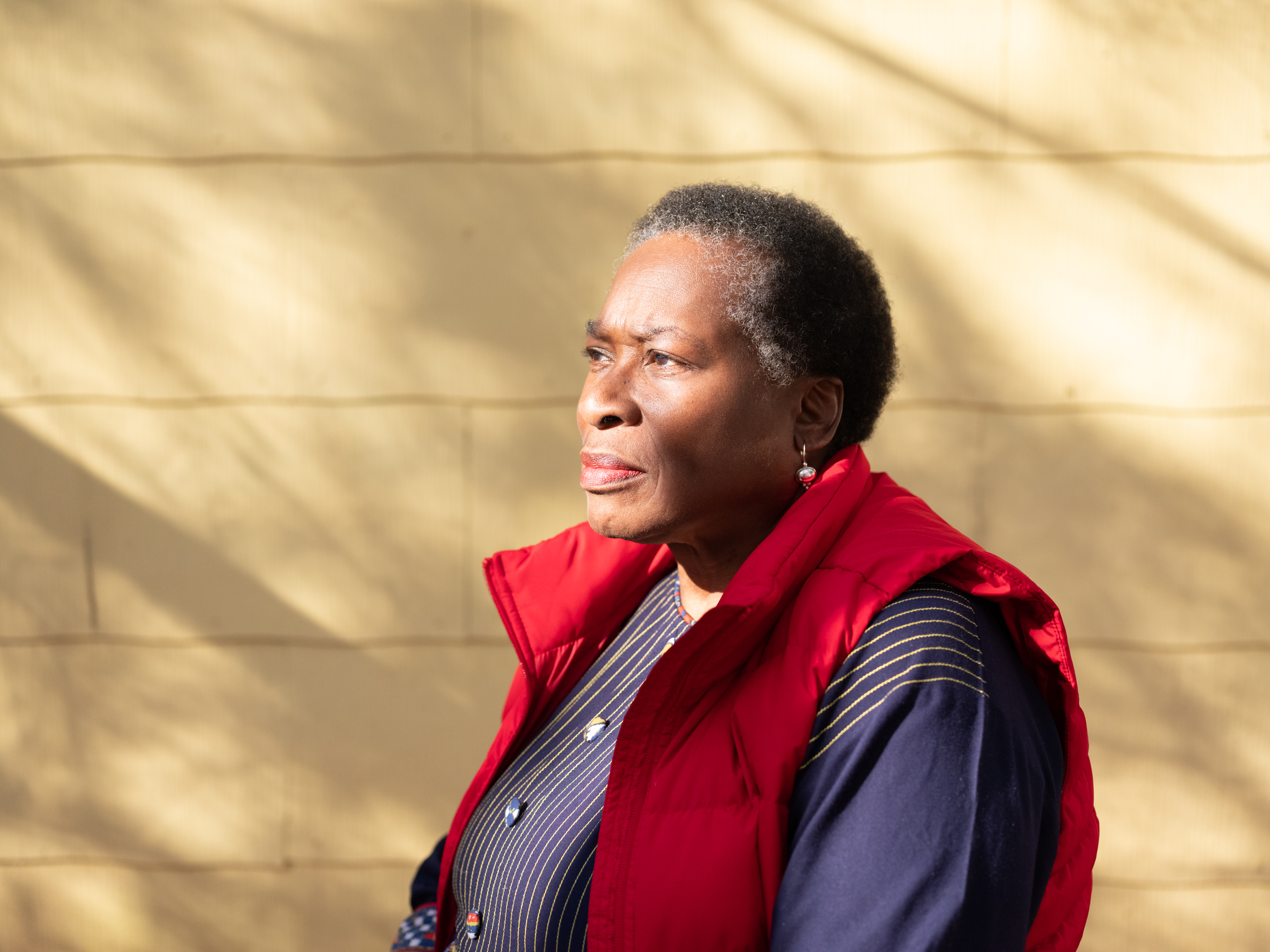 Lois is a Black woman with short, greying hair. She's standing outside in dappled sunlight, looking to the left, against a backdrop of a yellow home or other building. She's looking into the distance with a pensive expression.
