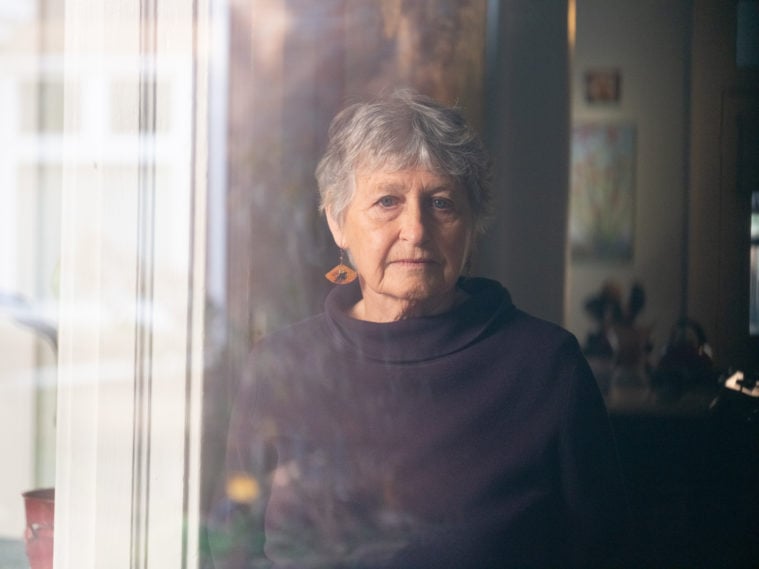 A white woman with short gray hair and large, dangly earrings, is visible through a residential window. She's gazing towards the viewer with a slight frown.