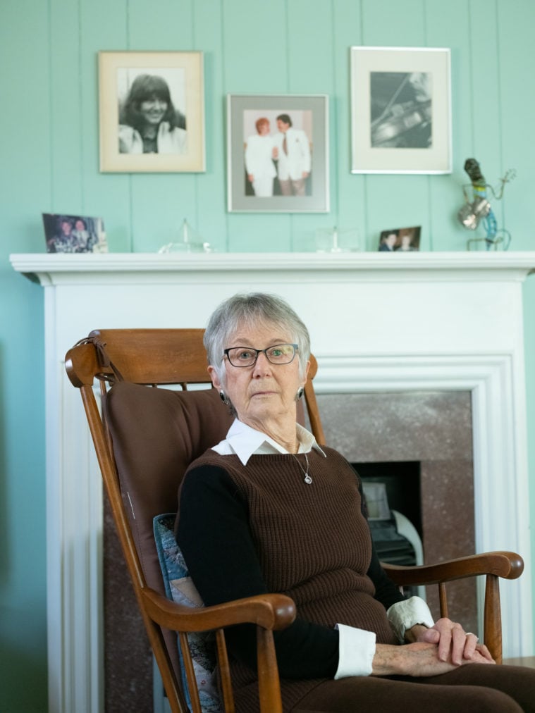 A white woman with short gray hair, wearing dark cats-eye glasses, gazes directly into the camera with a serious expression. She's sitting in an armchair with her arms folded. Behind her is a mantel decorated with knicknacks and family portraits.