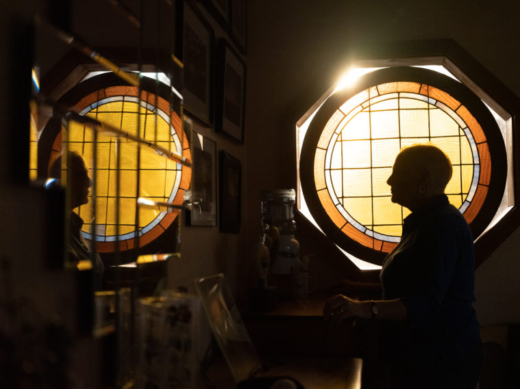 Sandra can only be seen in profile, looking to the left, with her profile reflected a second time in the mirrors attached to the wall of the darkened hallway. She's backlit in silhouette by a simple yellow and orange, circular stained glass window.