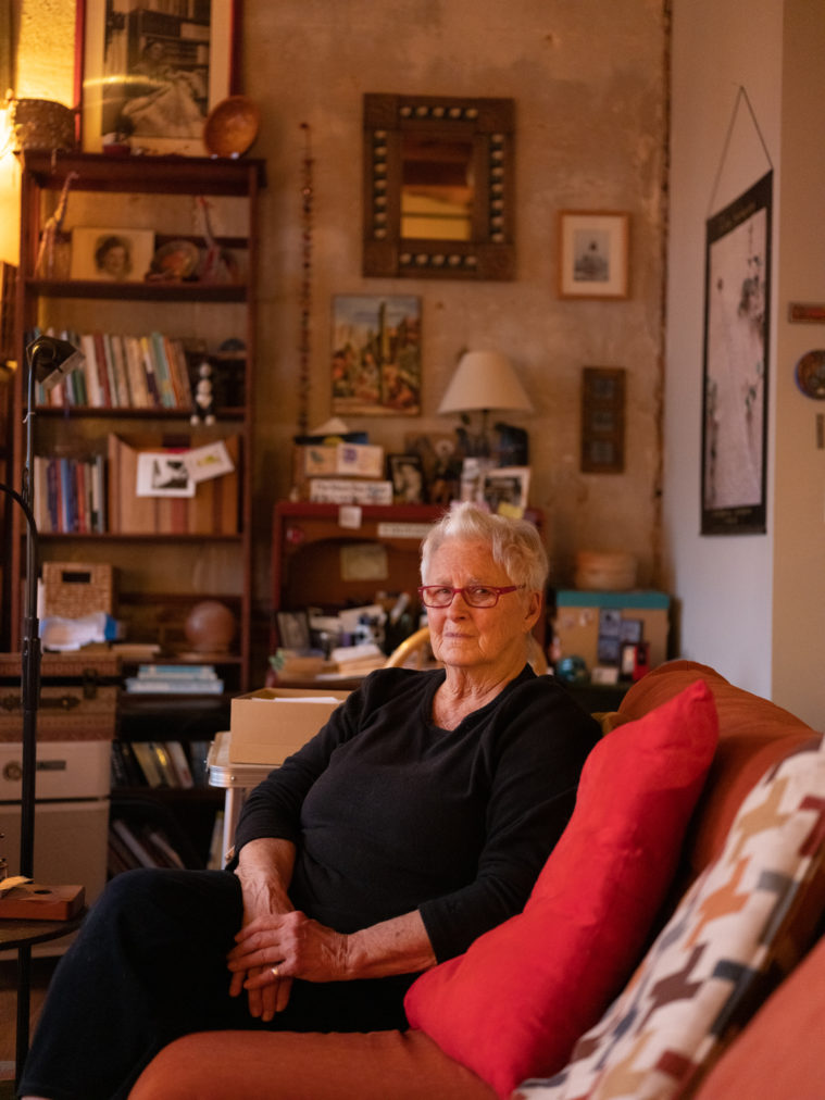 A white woman with short pale gray hair, sits with her arms folded in her lap on a couch. Behind her is her living room including a bookcase and cluttered desk.