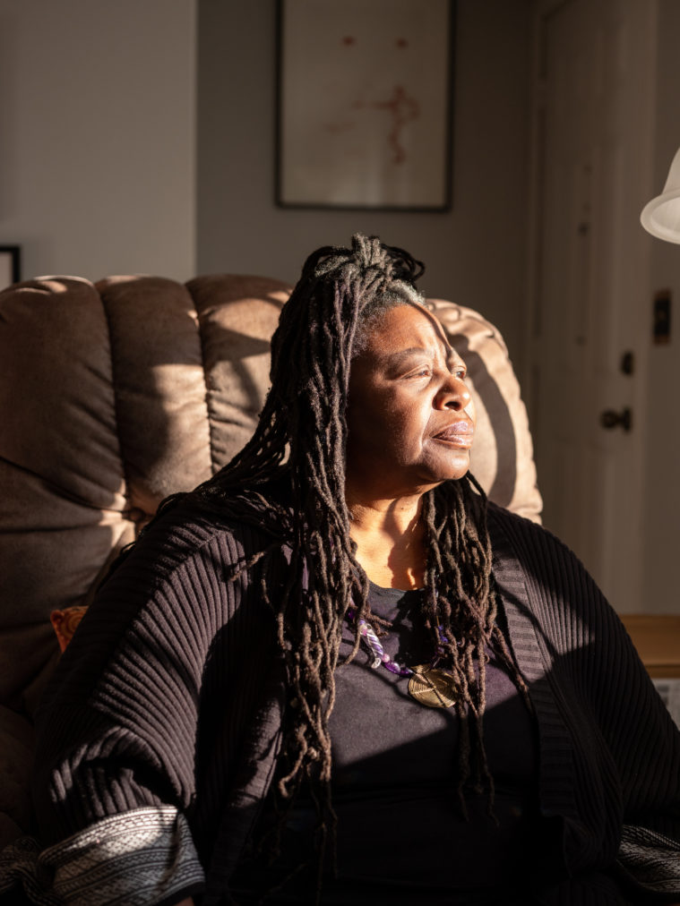 A Black woman with long, narrow locs past her shoulders sits in an easy chair. She's looking pensively to the right, with her face bathed in sunlight from a window out of frame.