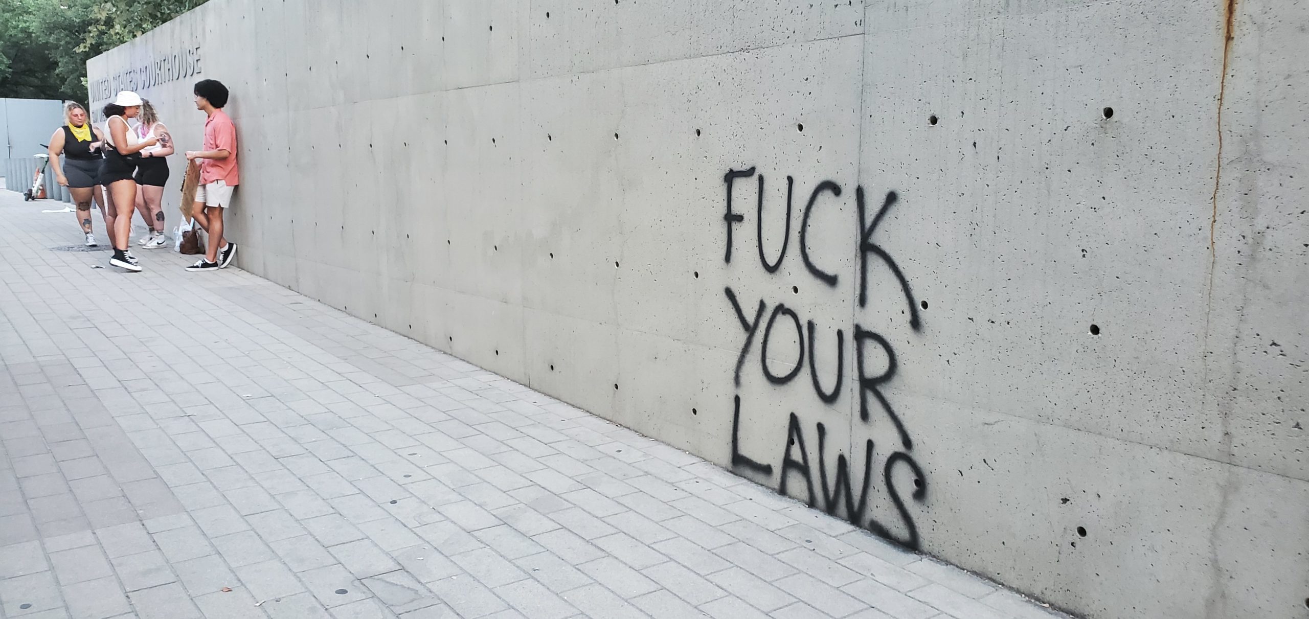 Graffiti on the side of the Federal Courthouse in Austin, Texas reads "Fuck Your Laws" in black spraypaint. In the background, a few activists gather.