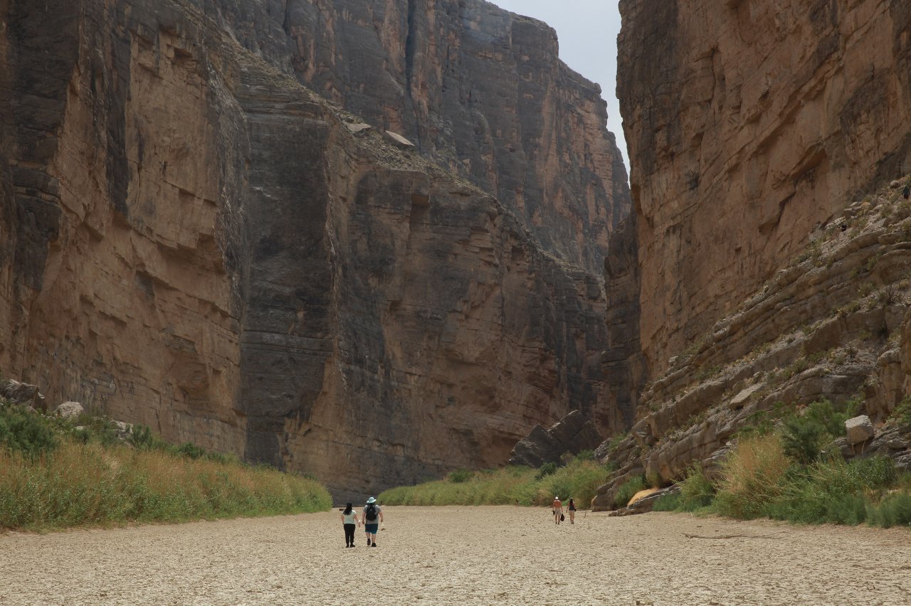 Tall canyon walls stretch to either side of the dry, cracked riverbed of the Rio Grande. Tewo people can be seen in the distance walking along it.
