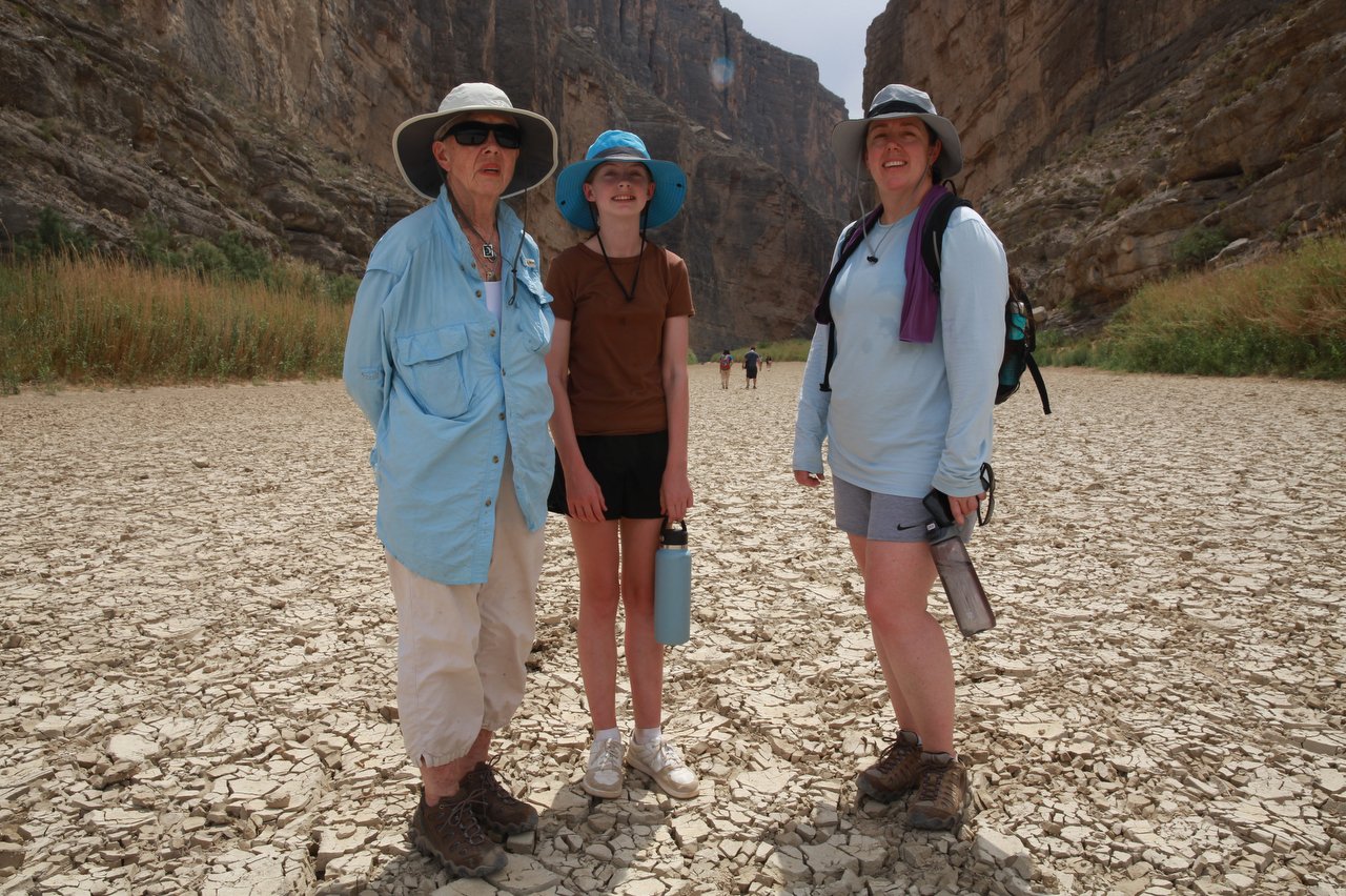 A trio of tourists in sun hats and hiking clothes stand on the dry, cracked riverbed of the Rio Grande.