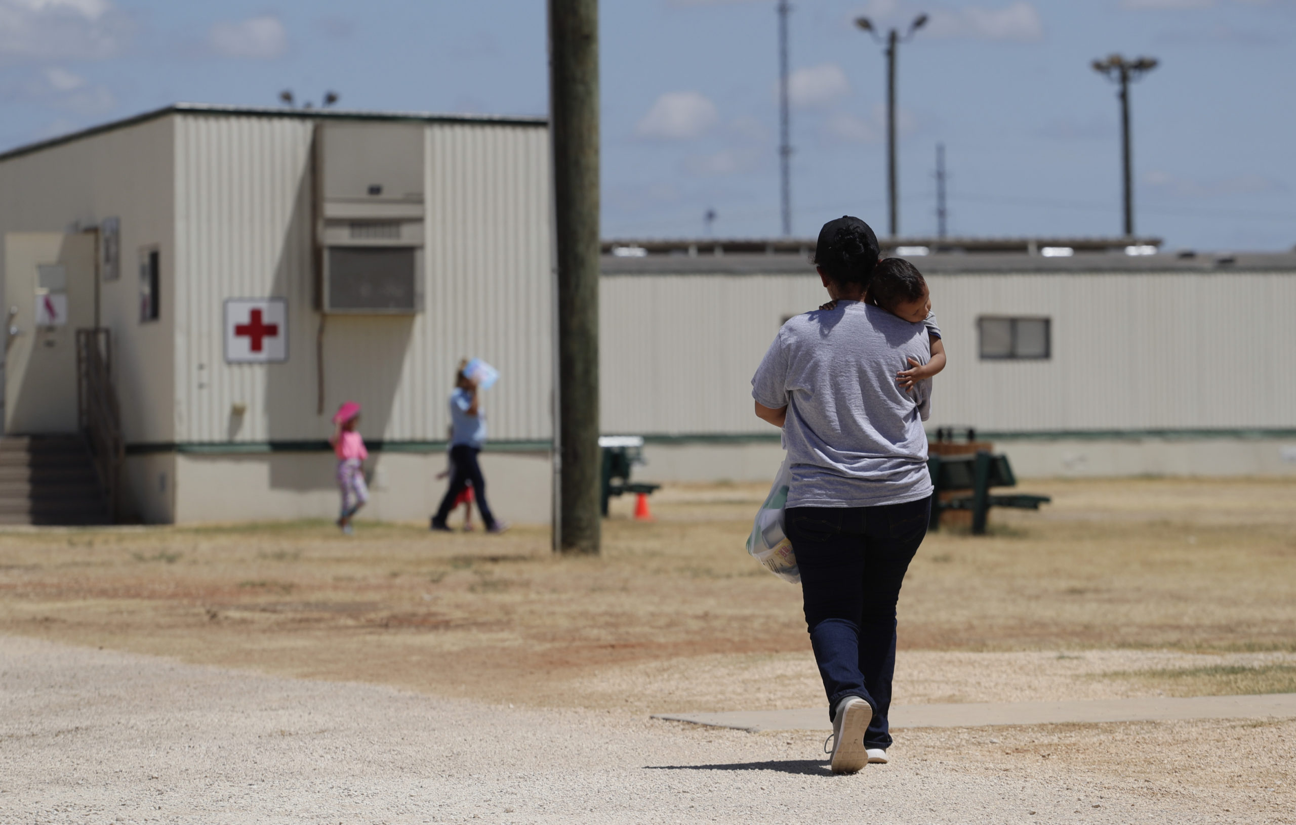 An immigrant mother holding a young child walks in a bare exercise yard at an ICE immigrant detention facility.