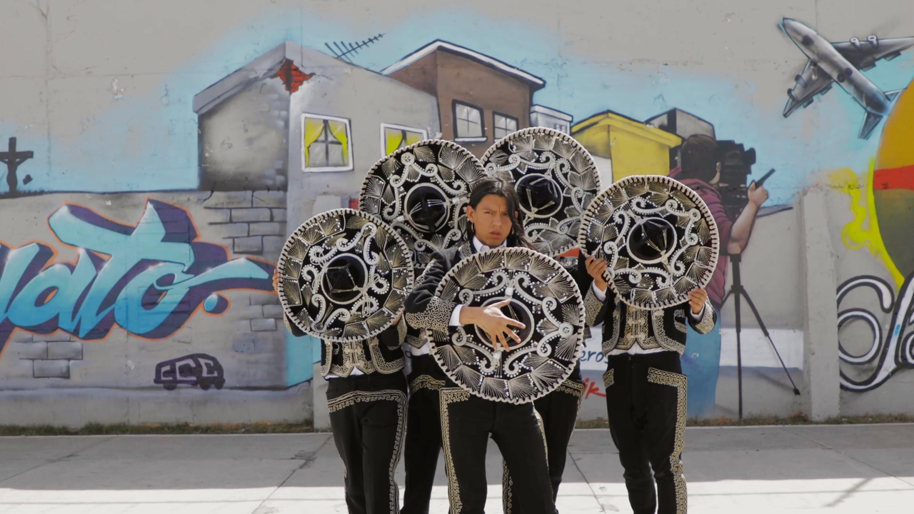 A group of five performers in black, gold and silver mariachi costumes perform in front of a graffiti-covered border wall.