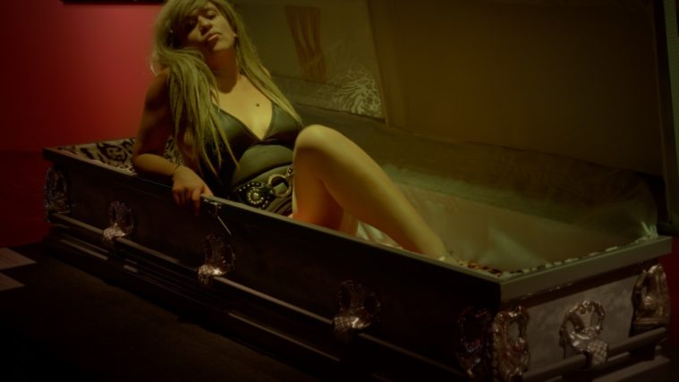 A long-haired performer in a dress poses like a model, lips pursed, while reclining in a coffin.