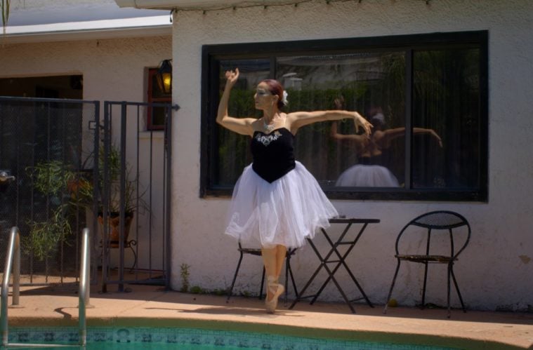 A ballerina in a white tulle skirt and black dress top dances on pointe by a swimming pool, near a sunbaked white building.