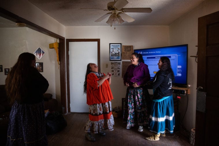 Three Native American women in a small cluster, standing in front of a TV in a living room, share laughter over a joke.