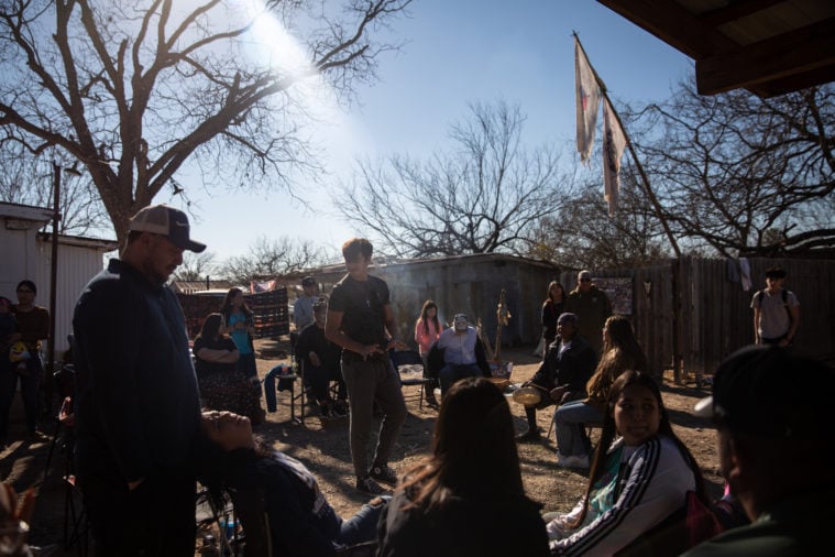 About a dozen people gather in a circle, standing or sitting on camp chairs, while some hold drums.