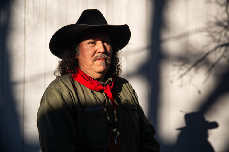 A distinguished looking Native American man gazes off to the right, wearing a black cowboy hat, red bandanna, dark green shirt and beaded necklace.