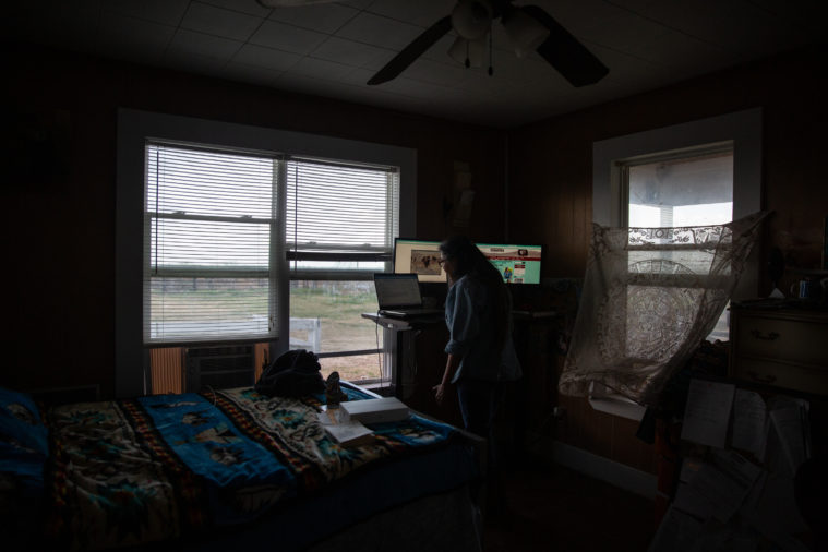 Lucille Contreras works on her laptop, with multiple monitors, at a standing desk in the corner of a dusky, windowed room.