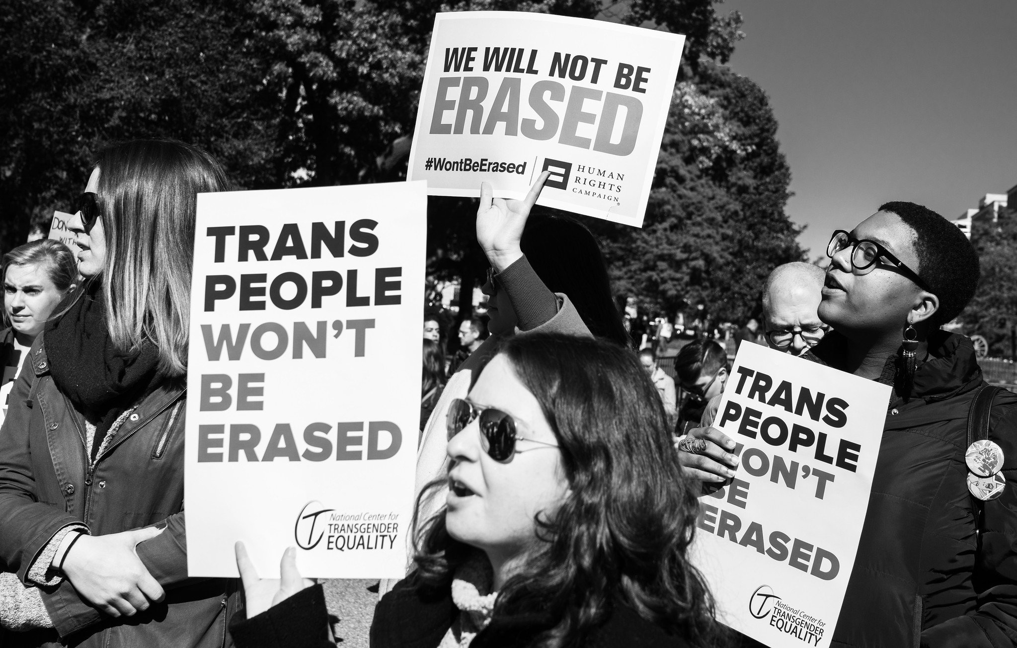 In a black and white photo, protesters hold signs demanding transgender rights, including "Trans People Won't Be Erased" and "We Will Not Be Erased".