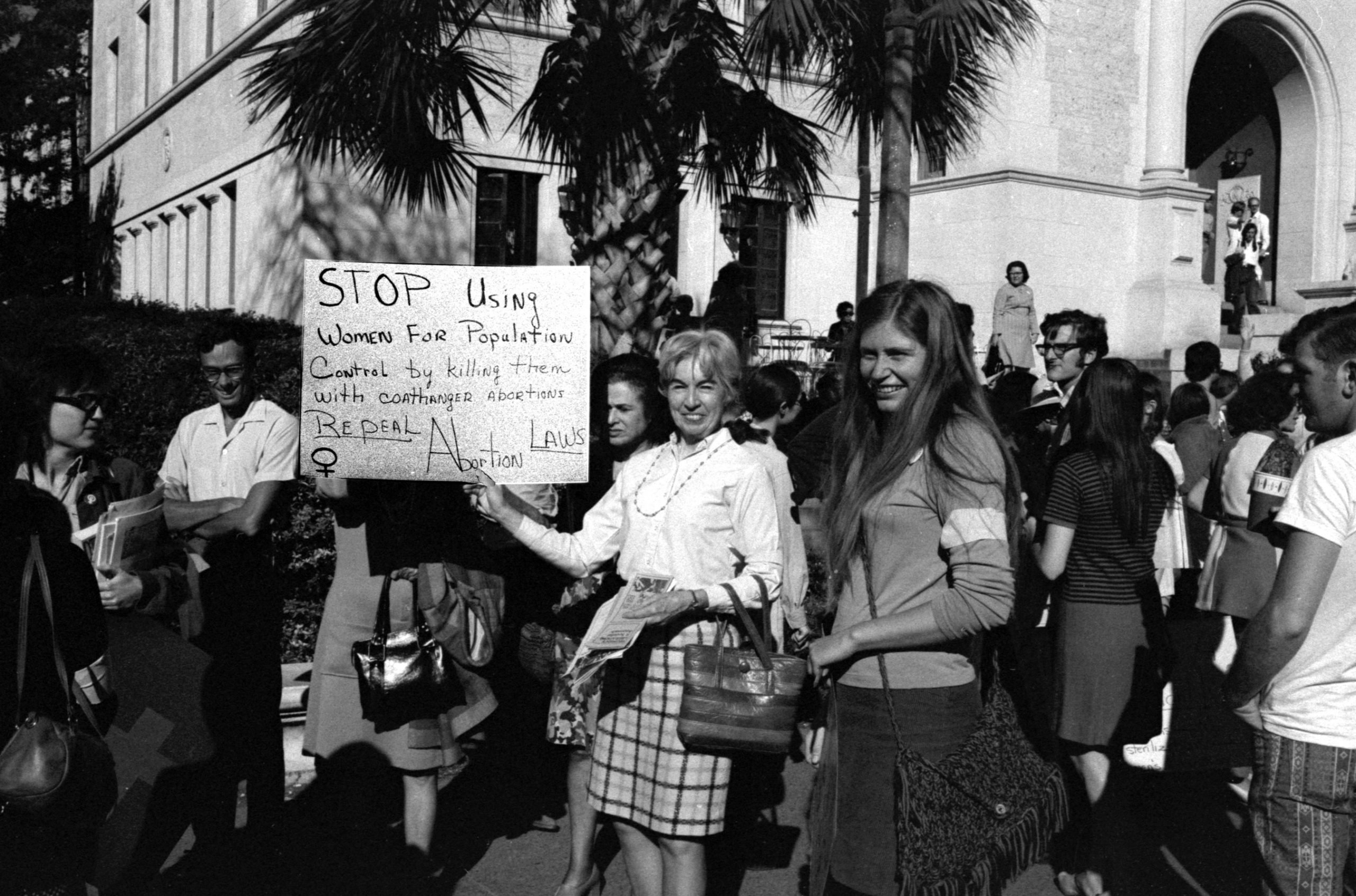 In a black & white photo, women demonstrate in favor of repealing abortion legislation in Texas. In the foreground, one woman holds a handwritten sign which reads "Stop using women for population control by killing them with coathanger abortions. Repeal Abortion Laws"