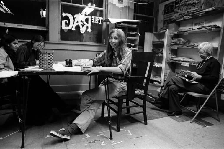 In a black and white photo, a group of journalists gather around a small table in a comfortably cluttered newspaper office. The logo of "The Rag" is visible in reverse through an outside window.