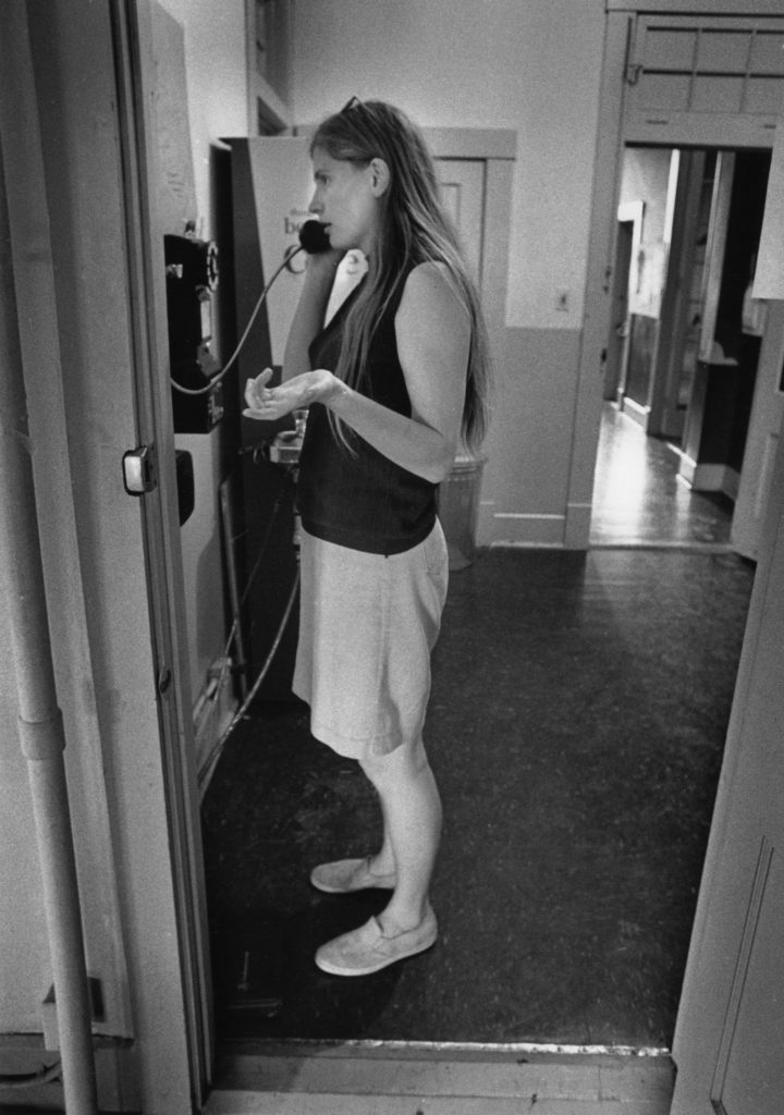In a black and white photo, a woman in shorts & a tank gestures conversationally as she speaks into a wall-mounted rotary phone in a hallway.