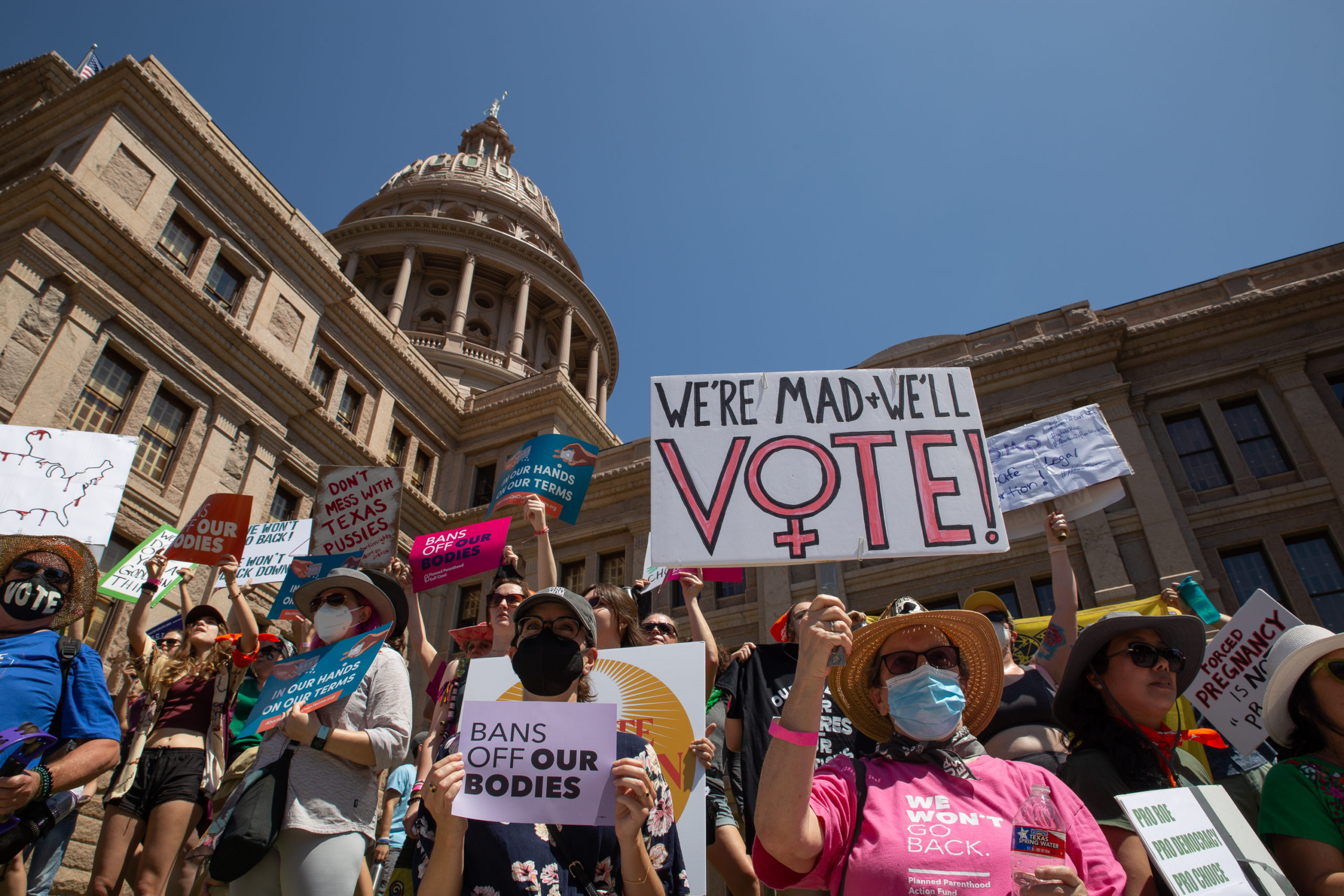 A group of reproductive rights proteters stand in front of the granite Texas capitol building, holding signs such as "Bans Off Our Bodies" and "We're Mad + We'll Vote!"