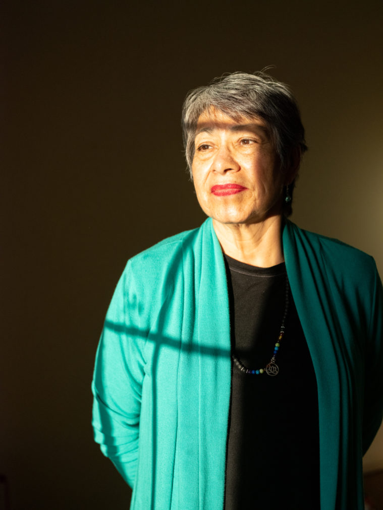 A Latina woman gazes pensively out a window, which is reflected in sunlight on her face. She's wearing red lipstick, a beaded necklace, and a turquoise sweater over a black shirt.