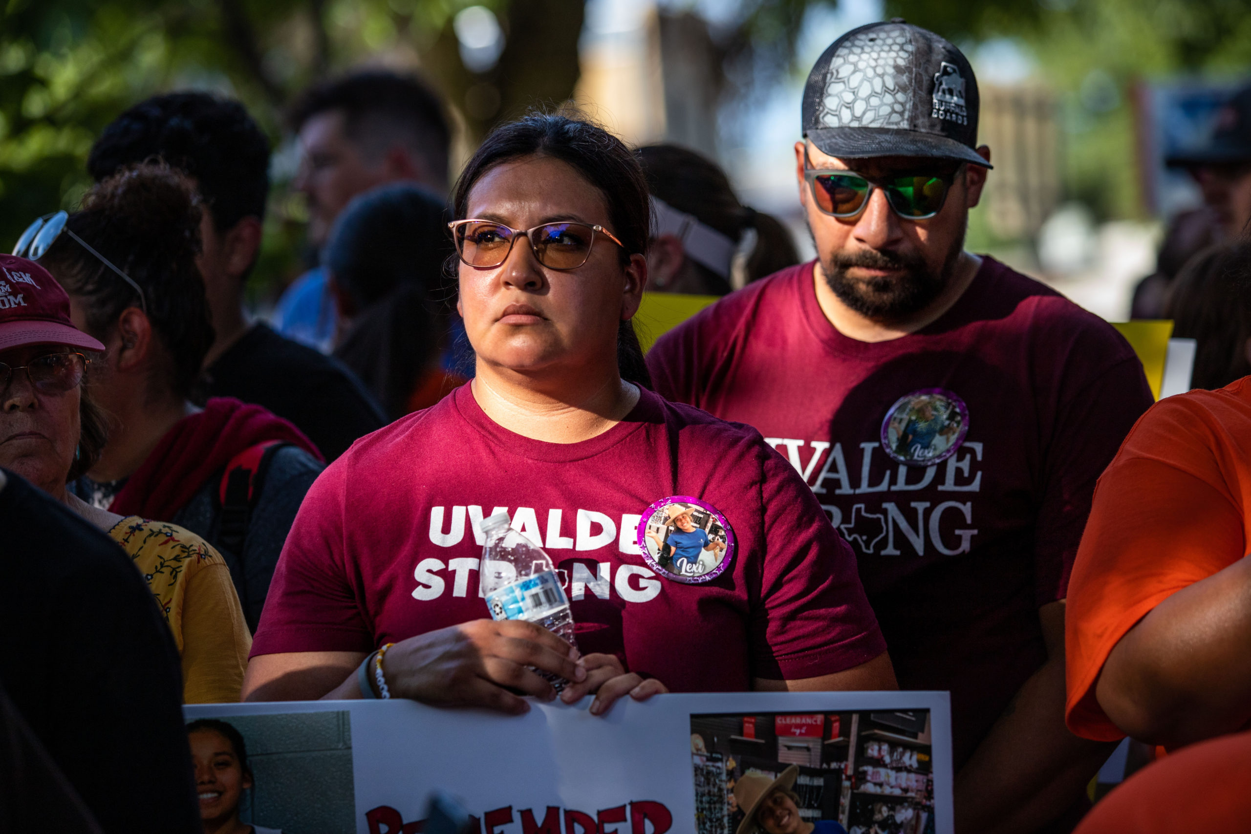 In a large crowd of people, a man and woman wear red Uvalde Strong t-shirt, their expressions sad.