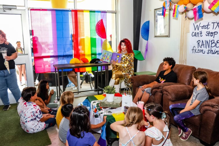 Children watch as a drag queen reads from a story book, wearing a deep red wig, gold sparkly dress and gold tights under white go-go boots.