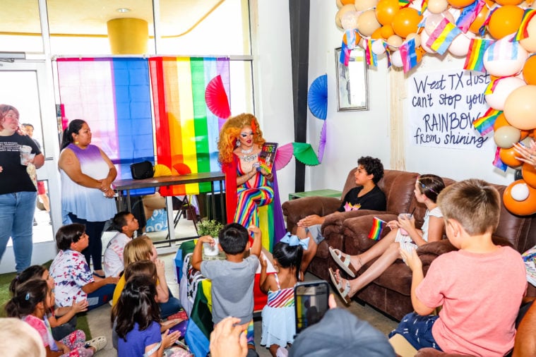 Over a dozen young children, along with some adults, are gathered to watch drag queen Miss Calvina read from a story book. Calvina is wearing silver jewelry, a red wig, and a rainbow dress and cape.