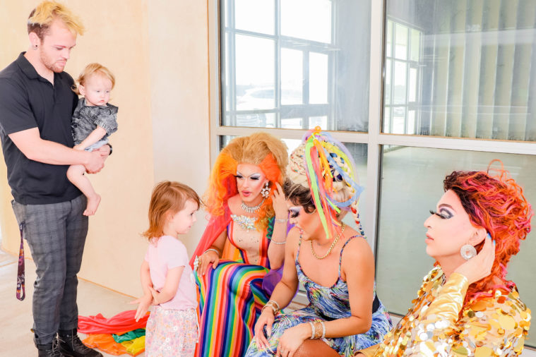 An adult holds a toddler as another young child in pink clothes speaks to the three drag queens outside the cafe'.