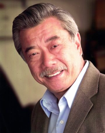 A headshot of Dana Lee, an older Chinese man with graying hair and moustache, wearing a button down shirt and suit jacket.