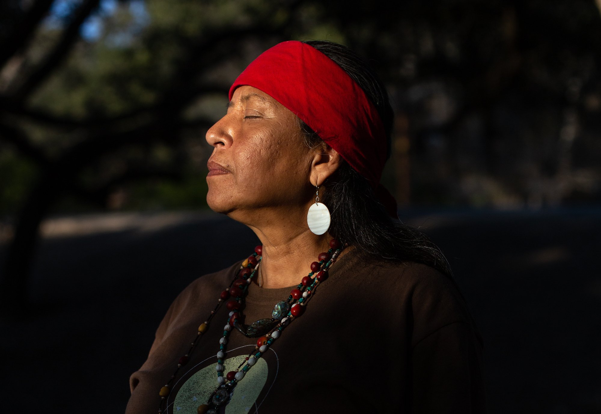 A Native American stands with a peaceful expression, eyes closed, wearing a red head covering and beaded necklace.