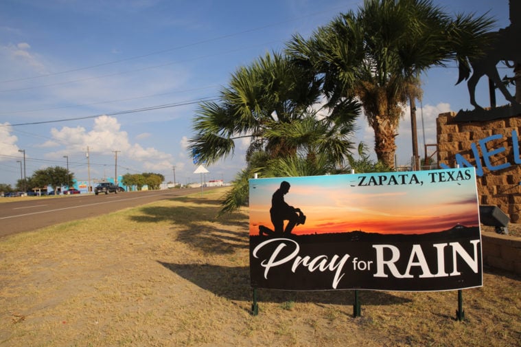 A sign int he dry dirt near a roadway shows a person kneeling, holding a cowboy hat, at sunset, with the text, "Zapata, Pray for Rain".
