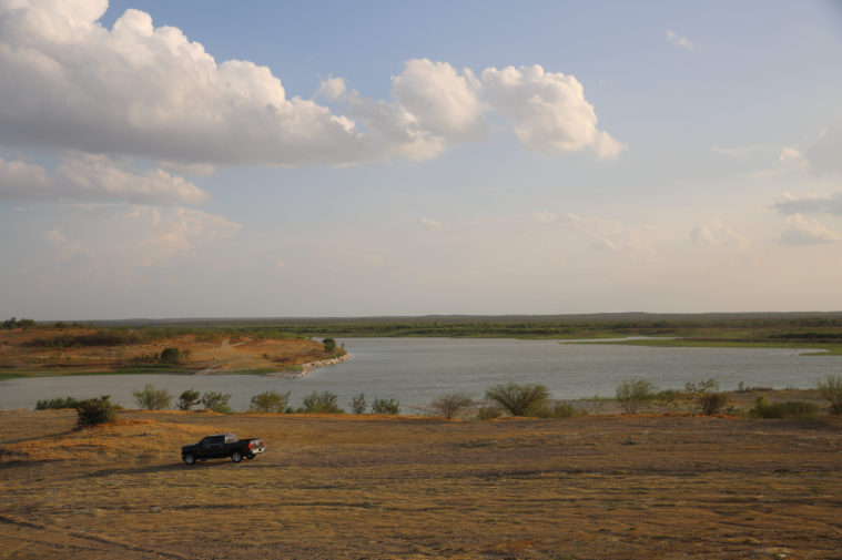 A lakebed that is almost dry, with a truck driving by in the foreground, under a big, partly cloudy sky.