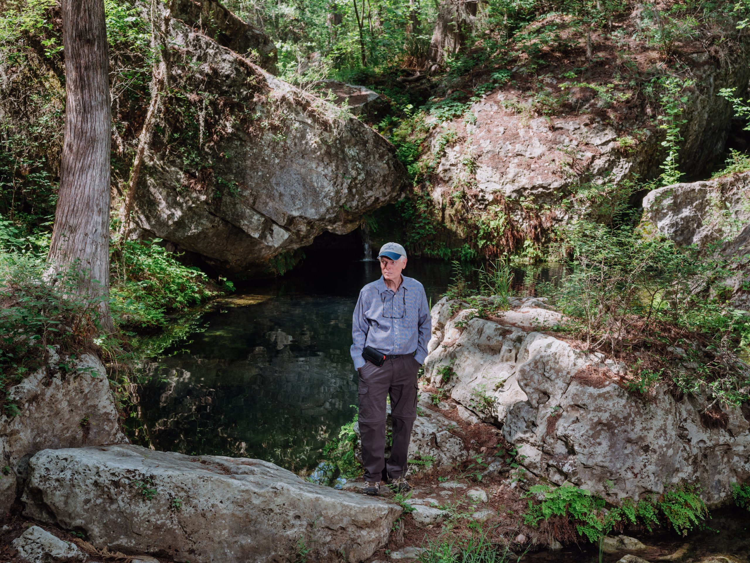 Lew Adams, an older man in a ballcap, denim shirt and dark slacks, stands among natural splendor at a small spring surrounded by a cave and green foliage.