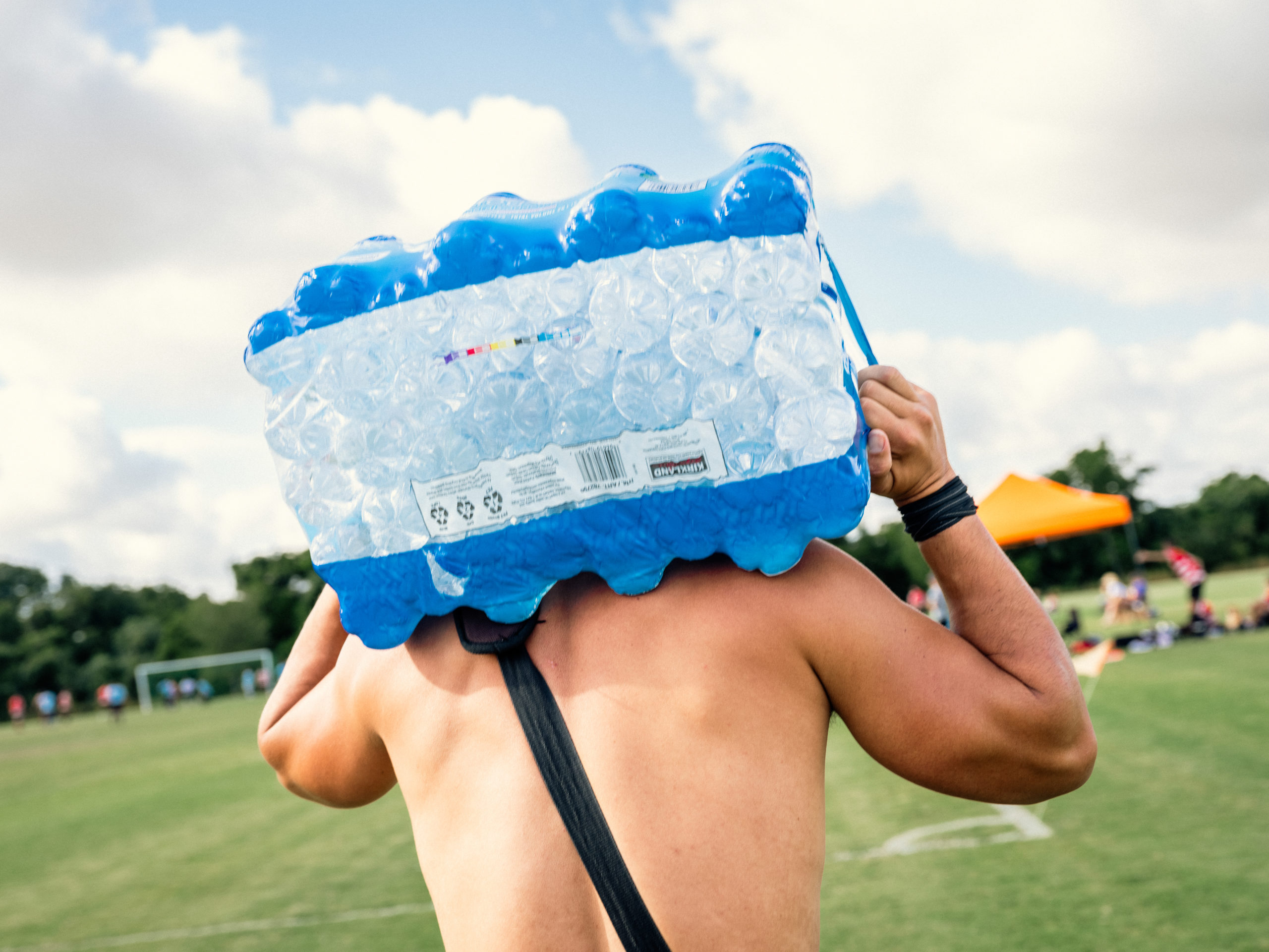 A shitless man carries a large, plastic-wrapped case of bottled water across a green field.