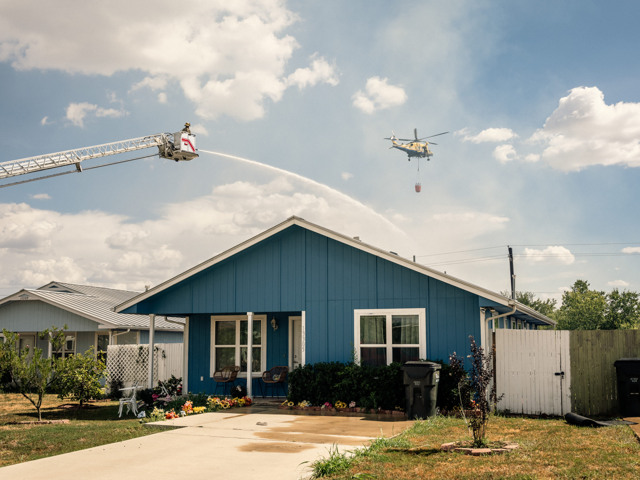 A fire fighter on an extended ladder, and a helicopter are used to spray water onto a brush fire behind a residential home.
