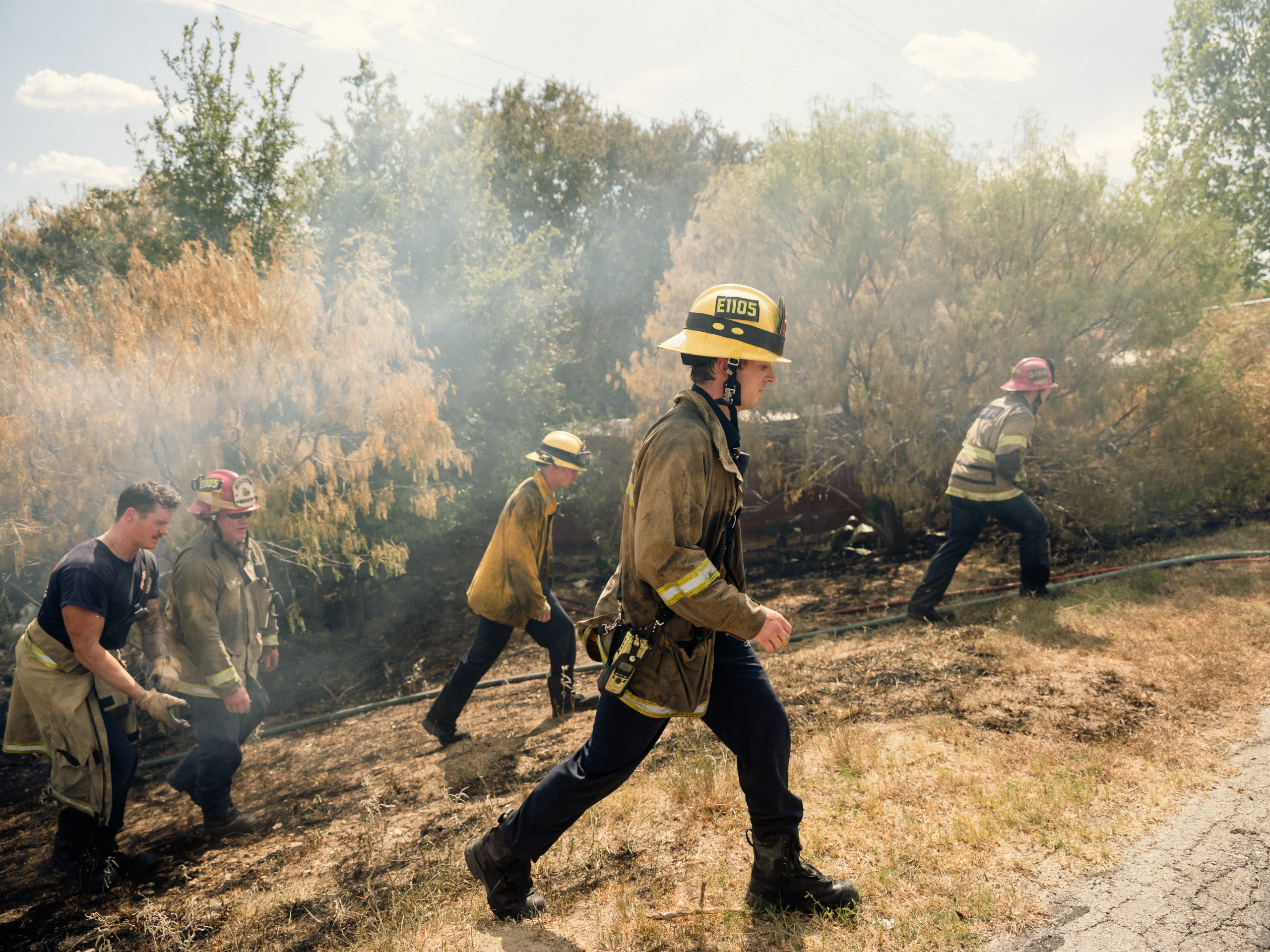 A crew of firefighters in uniform carry their gear up a steep, smoky hill.