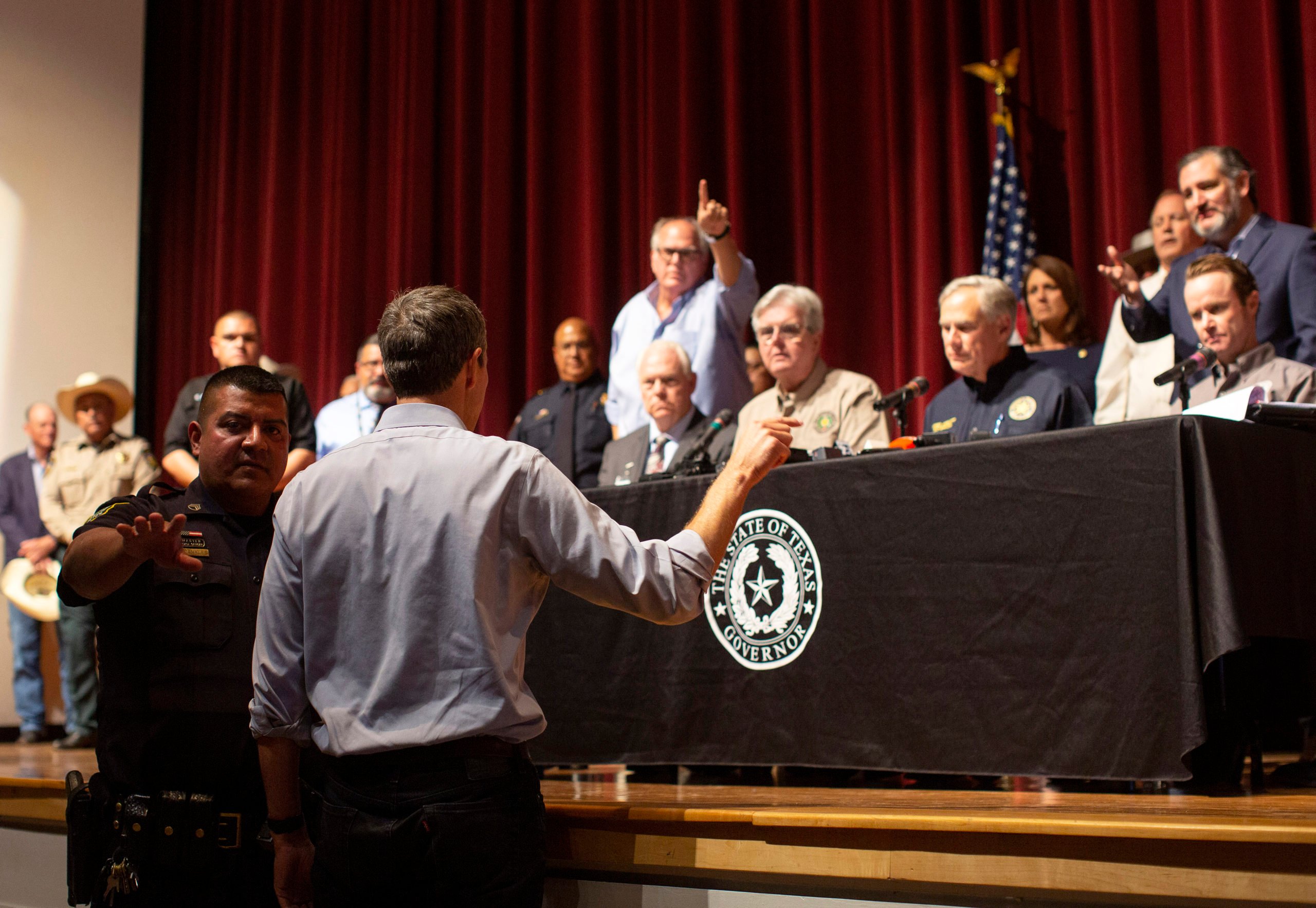 A police officer attempts to restrain Beto O'Rourke as he confronts Greg Abbott, who is seated on a dais. Behind Greg Abbott, the Uvalde mayor points angrily at O'Rourke.