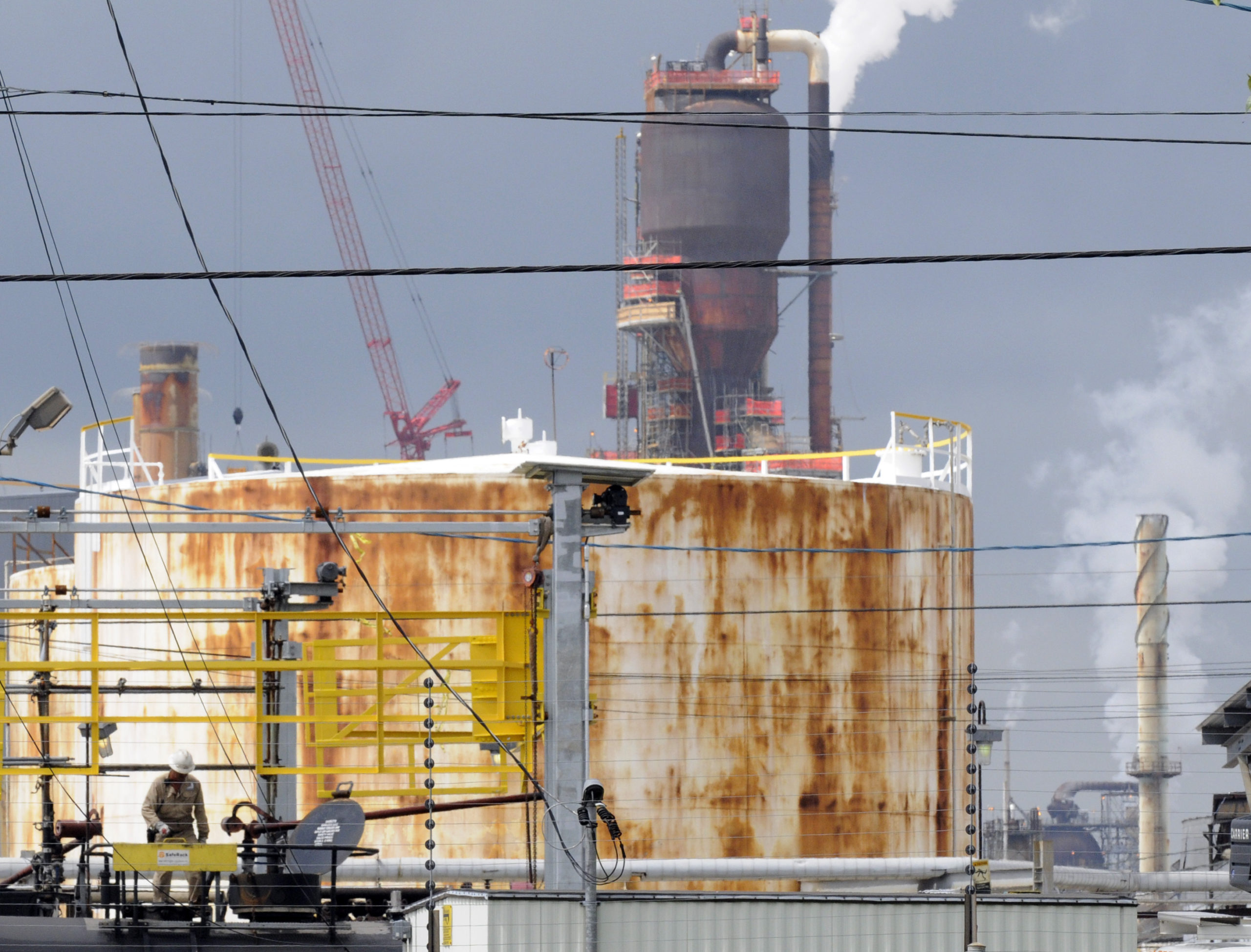 A worker can be seen in the foreground, while in the background is a chemical tank and smokestack of a refinery, with a small white plume emerging.