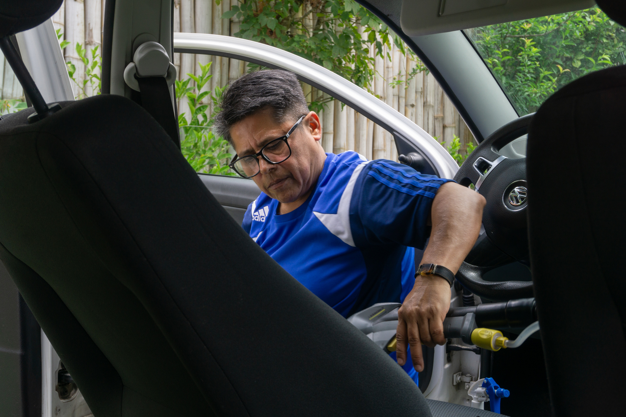 A Mexican person with graying hair works on the interior of a car. He can be seen from the waist up, wearing a blue Adidas shirt.