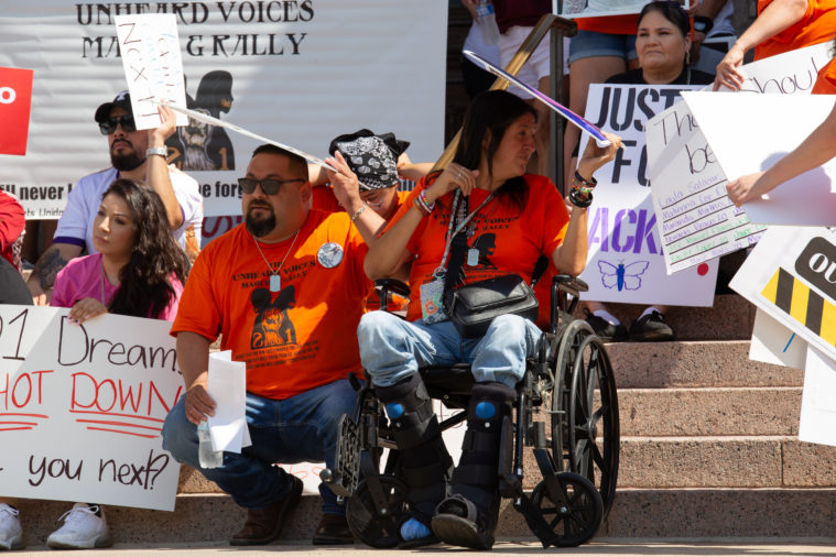 Family members from Uvalde use protest signs to create shade from sun.