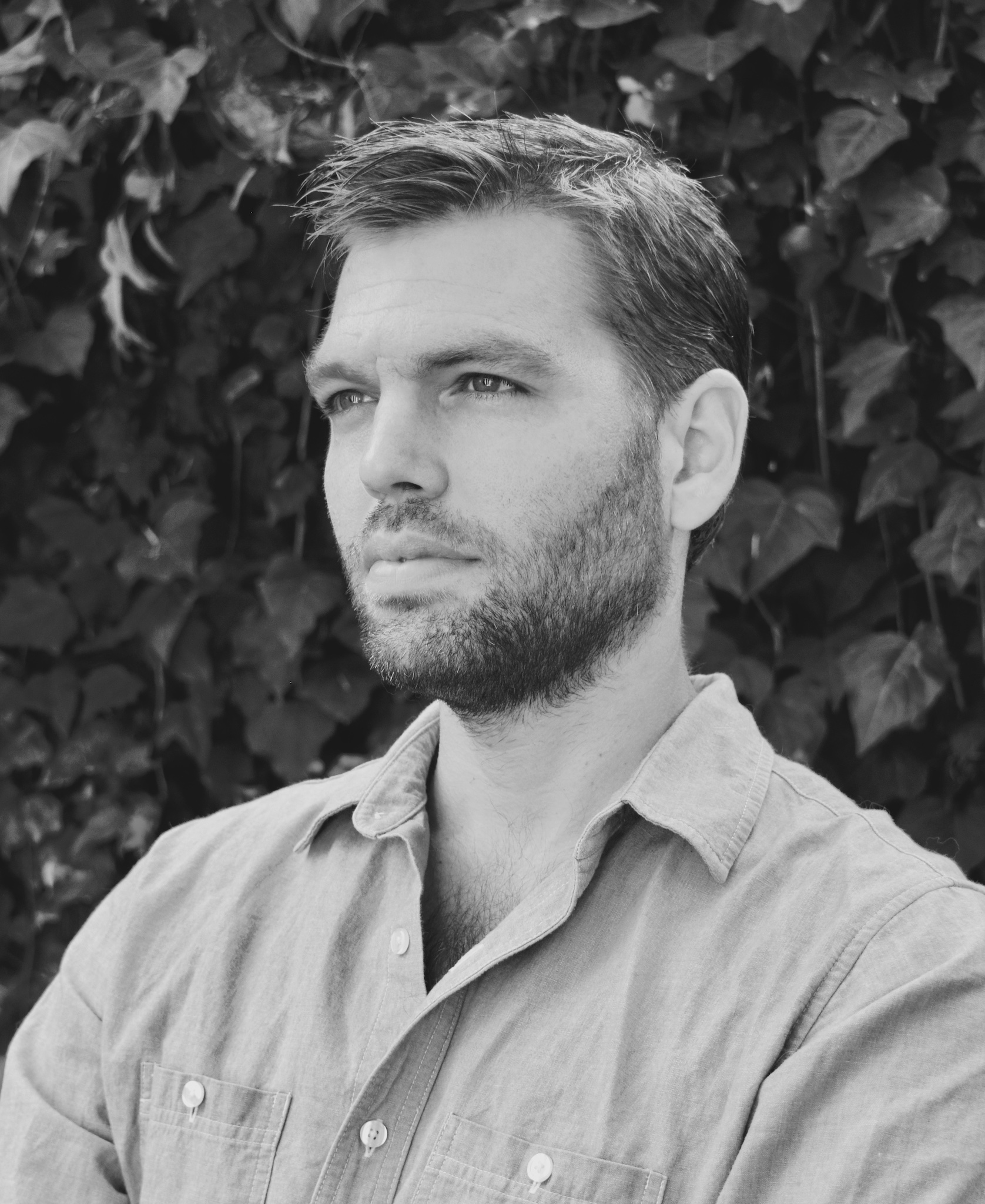 Kirk Johnson, a white man with short hair and a close-cropped beard, poses in a black and white photo. he's wearing a button-down and standing in front of an ivy covered wall.